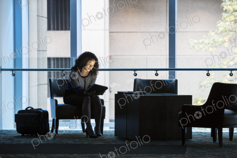 A black businesswoman sitting in front of windows in a darkened lobby area of a…