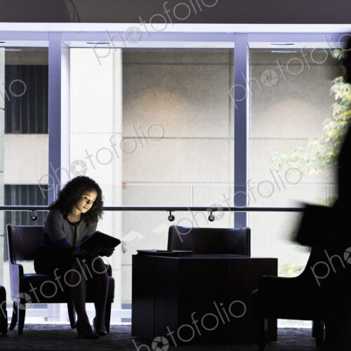 A black businesswoman sitting in front of windows in a darkened lobby area of a… A black businesswoman sitting in front of windows in a darkened lobby area of a…
