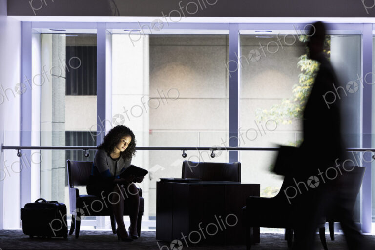 A black businesswoman sitting in front of windows in a darkened lobby area of a…
