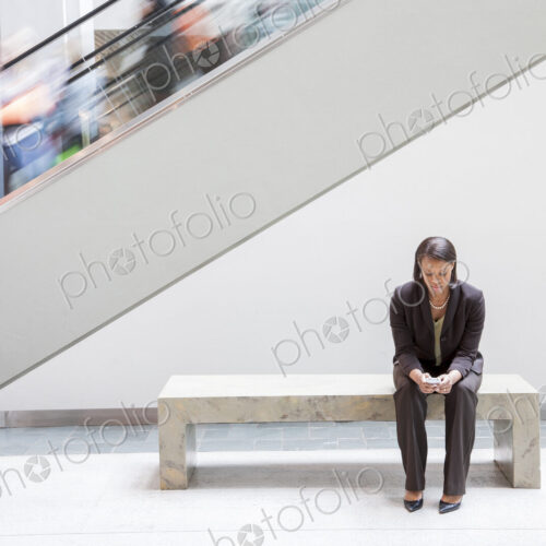 A black businesswoman texting on a cell phone while sitting next to an escalator.