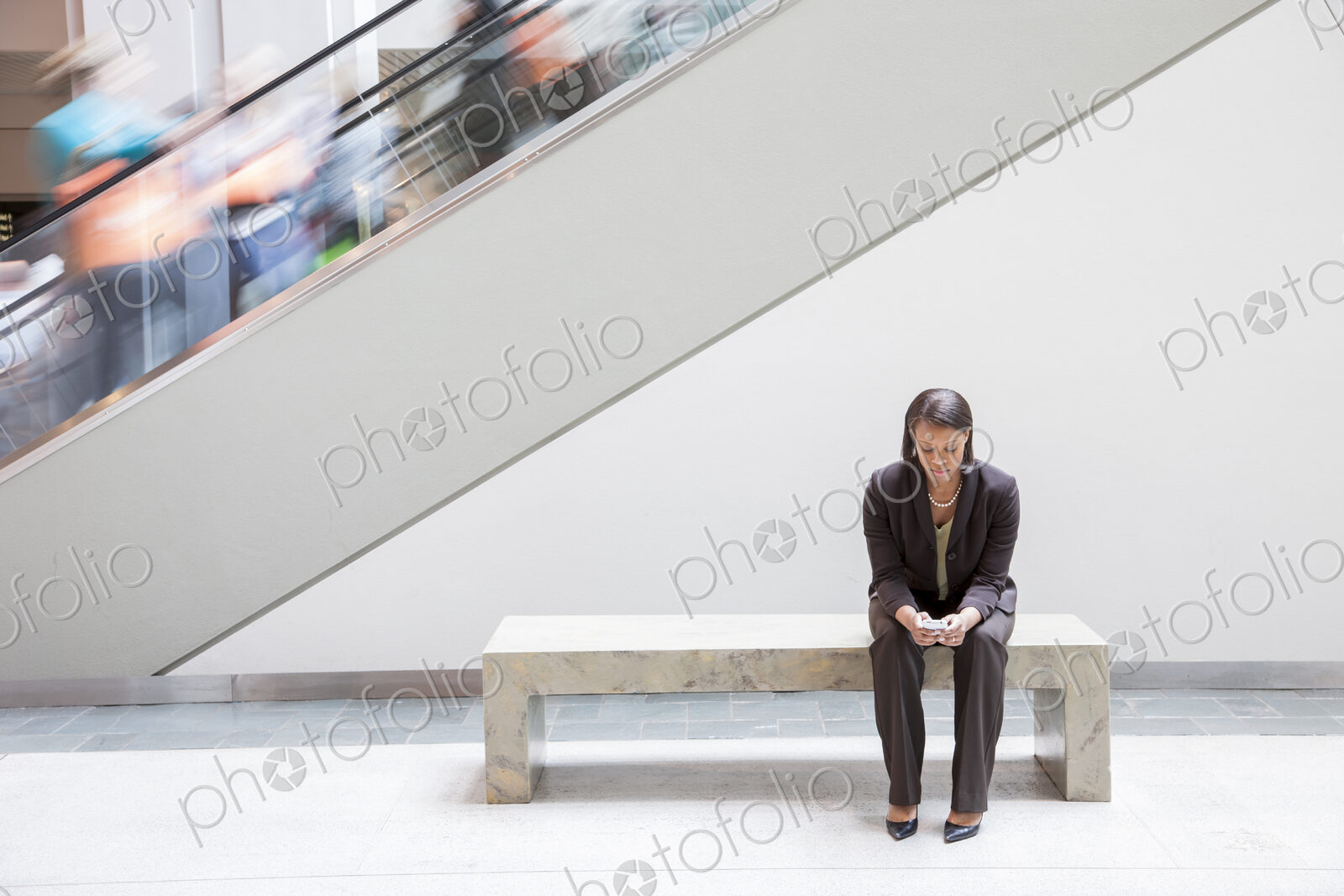 A black businesswoman texting on a cell phone while sitting next to an escalator.