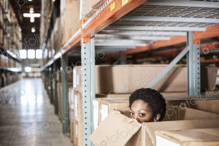 A black female worker hiding inside a cardboard box in a distribution warehouse.