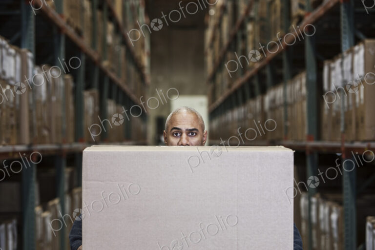 A black warehouse worker hidden behind a cardboard box in a distribution warehouse.