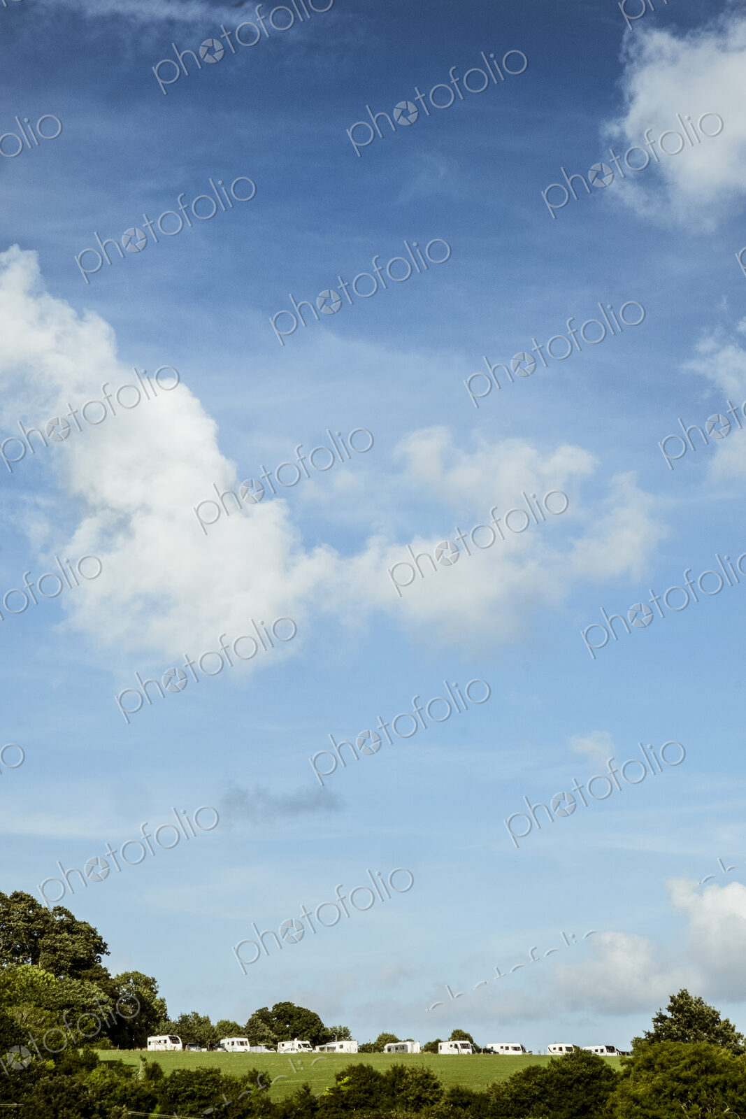 A blue sky with light cloud, and a row of caravans parked in a green field. Trees.