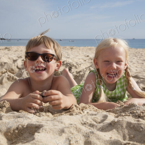 A boy and girl lying on their stomachs on the sand, laughing and looking at the camera. A boy and girl lying on their stomachs on the sand, laughing and looking at the camera.