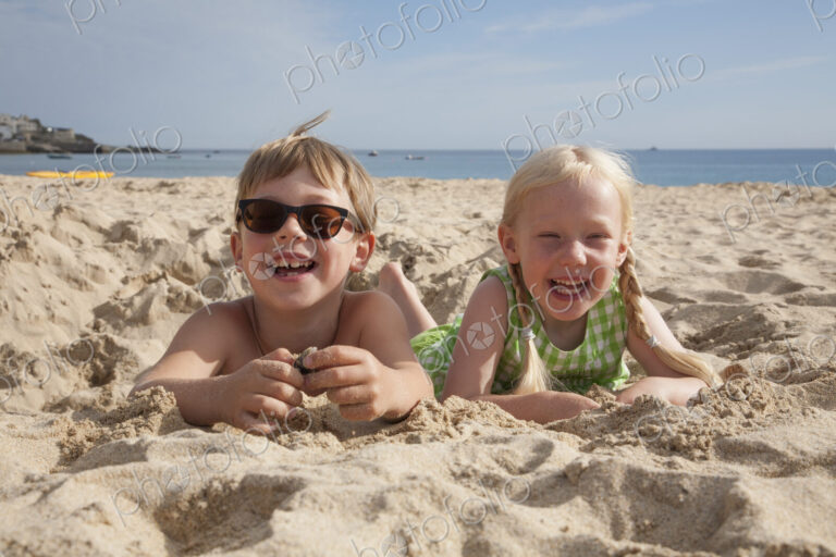 A boy and girl lying on their stomachs on the sand, laughing and looking at the camera.