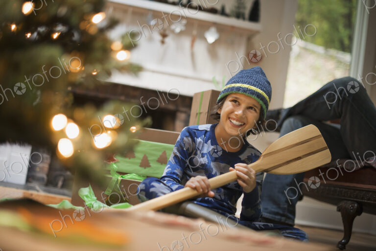 A boy by a Christmas tree unwrapping a gift, a wooden oar.