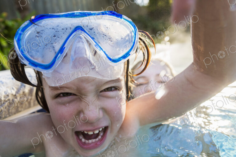 A boy in goggles laughing at the camera, in a warm swimming pool with his sister.