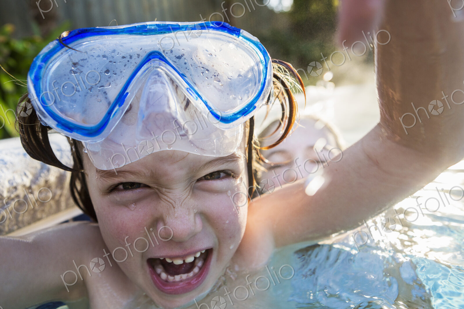 A boy in goggles laughing at the camera, in a warm swimming pool with his sister.