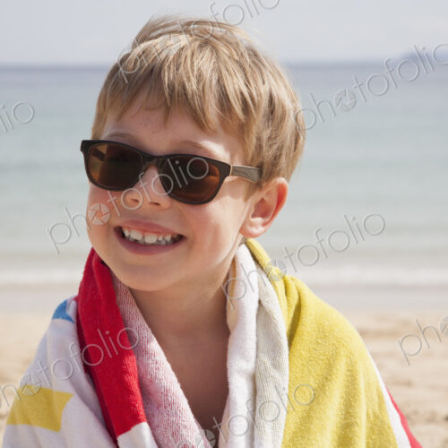 A boy in sunglasses on the beach, with a towel around his shoulders.