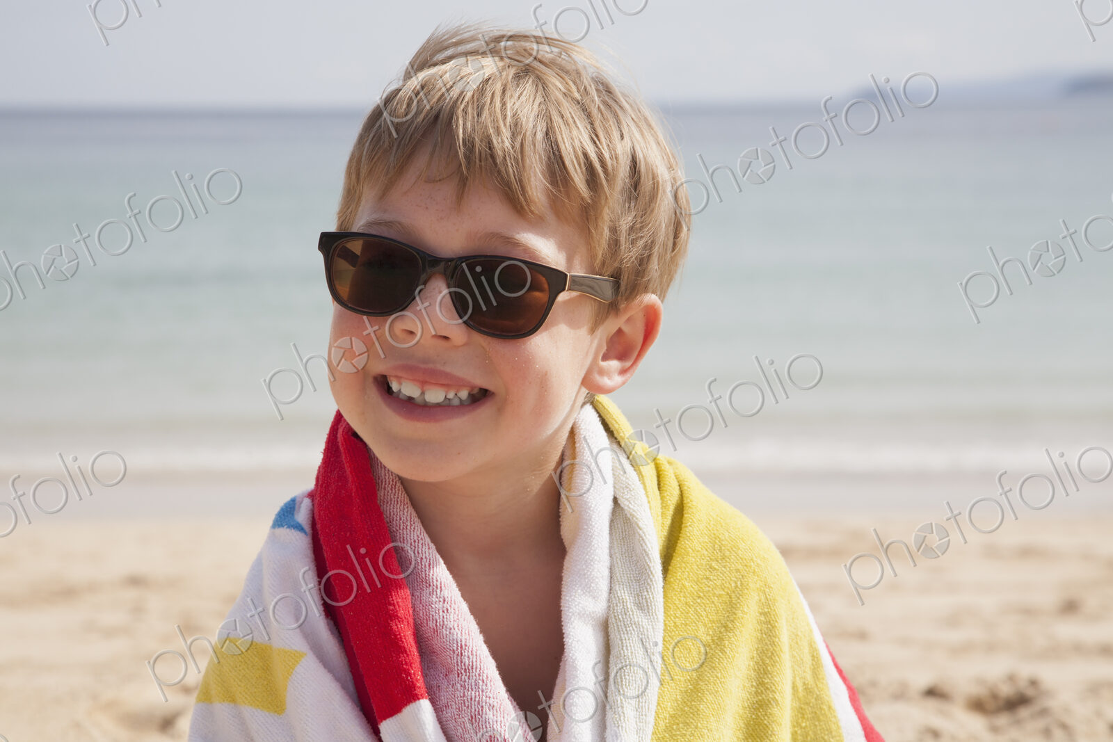 A boy in sunglasses on the beach, with a towel around his shoulders.