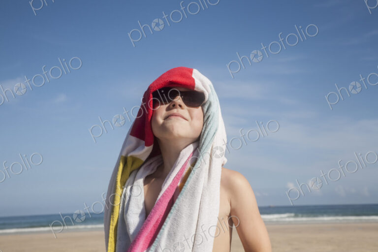 A boy in sunglasses with a towel over his head.
