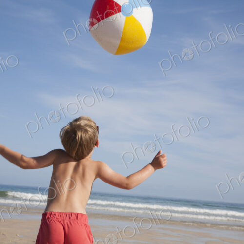 A boy in swimming trunks on the beach, with a large beach ball in the air above him.