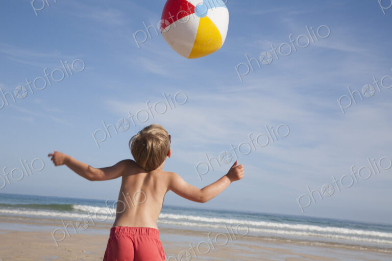 A boy in swimming trunks on the beach, with a large beach ball in the air above him.
