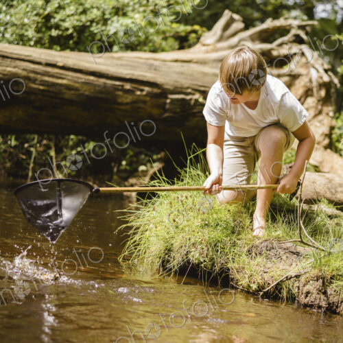 A boy kneeling by the river bank, leaning over and using a small fishing net. A boy kneeling by the river bank, leaning over and using a small fishing net.