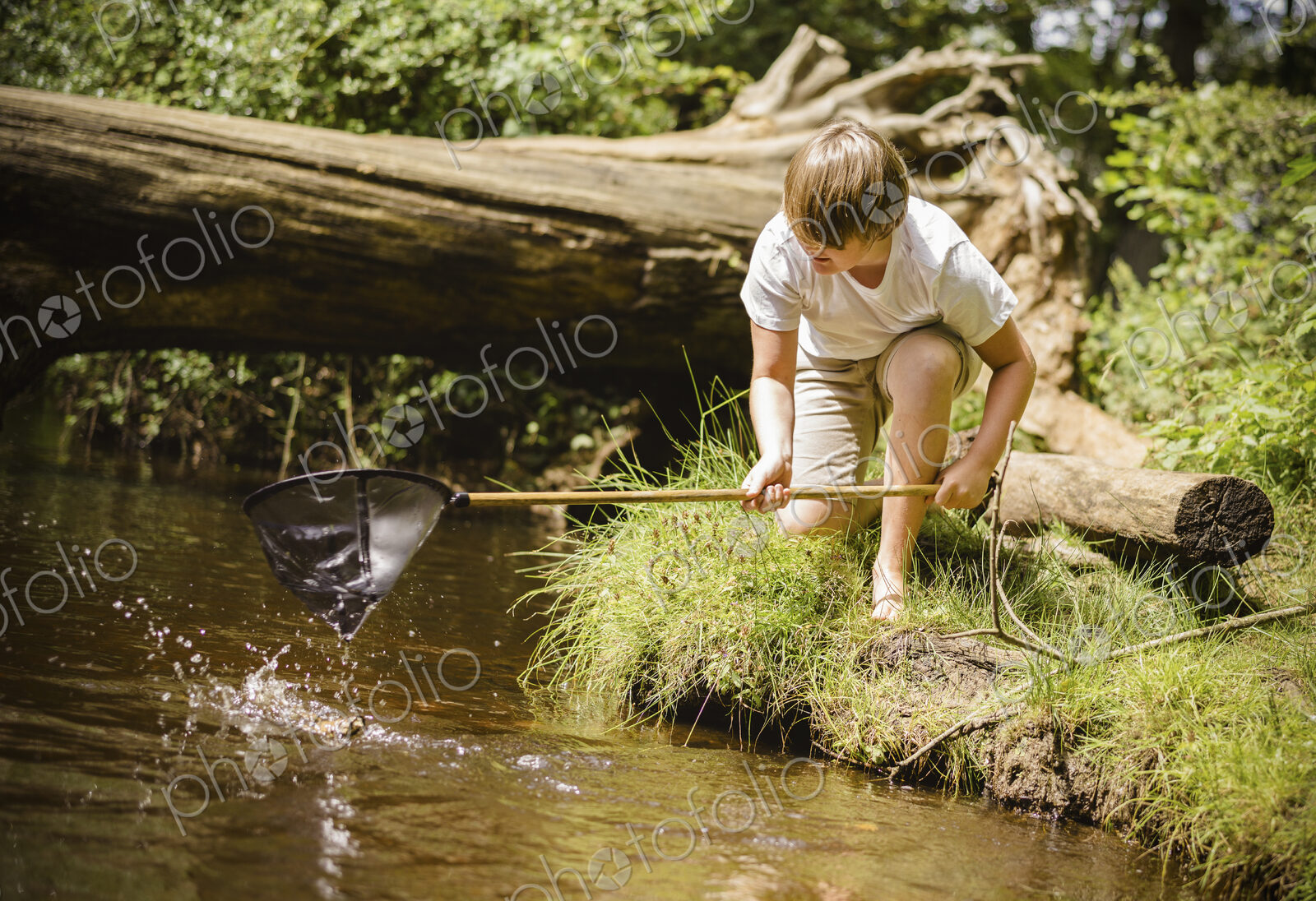A boy kneeling by the river bank, leaning over and using a small fishing net.