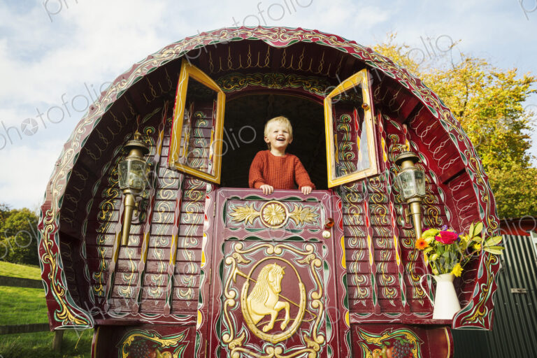 A boy leaning out of the open shutters at the rear of a bow top gypsy caravan.