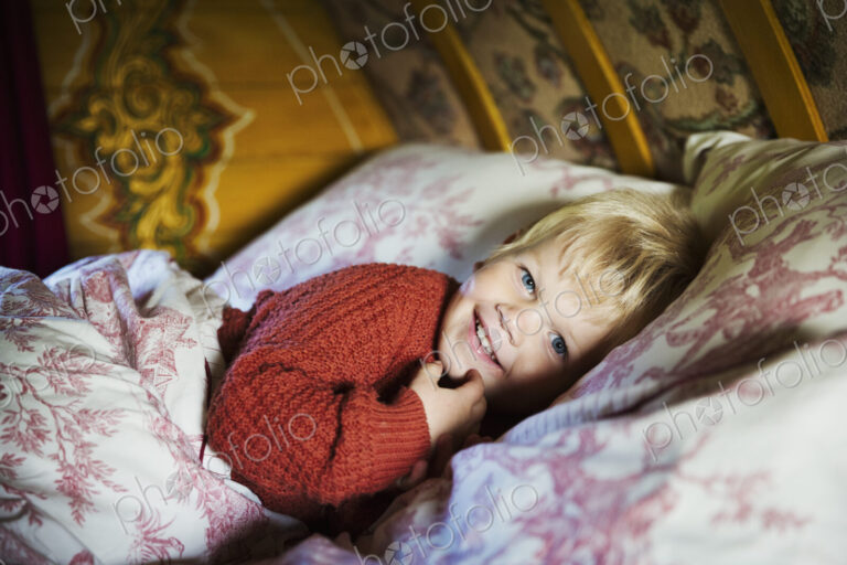 A boy lying in bed under a curved bow top roof of a gypsy caravan, looking at the camera.