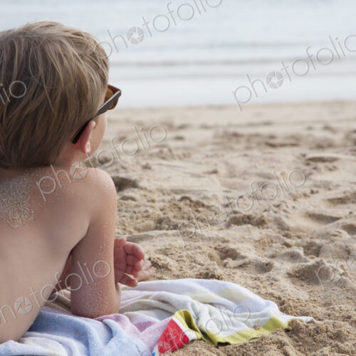 A boy lying on his front on the sand looking out to sea.