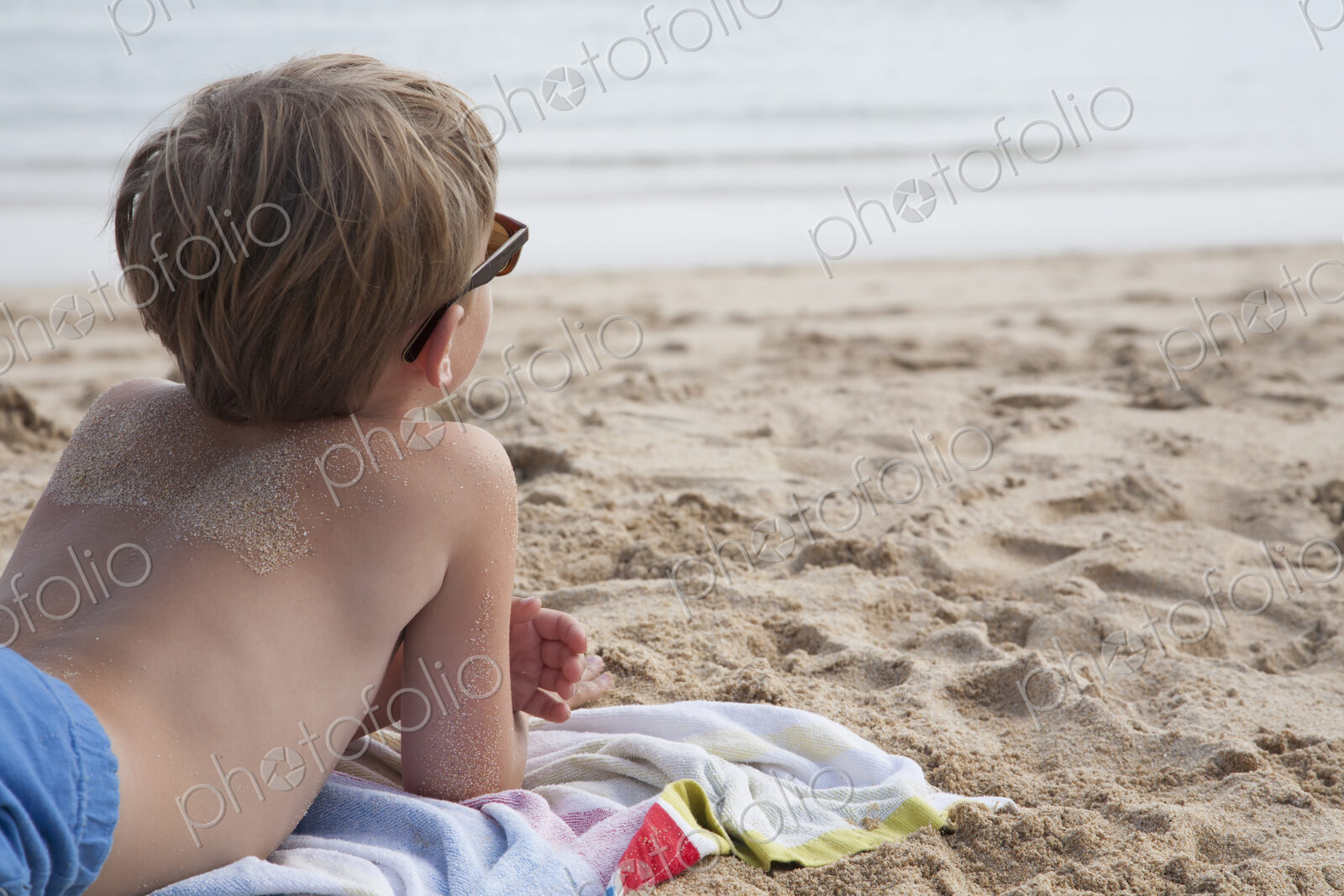 A boy lying on his front on the sand looking out to sea.