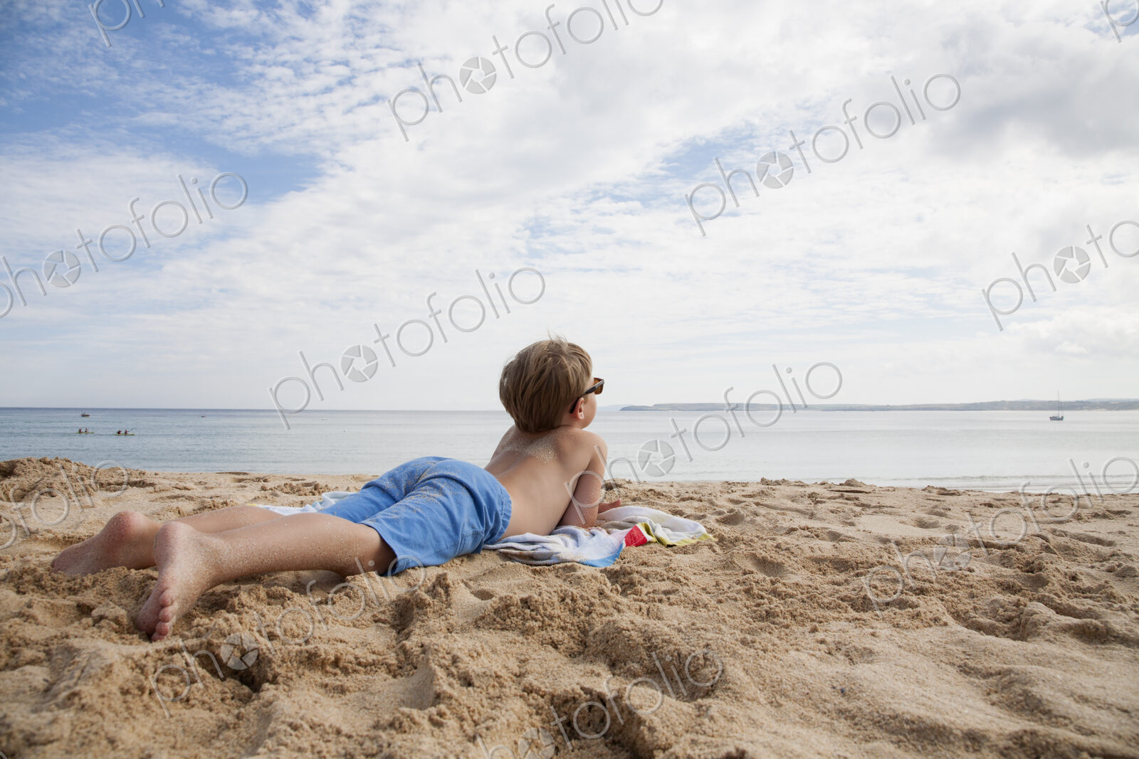 A boy lying on his front on the sand looking out to sea.