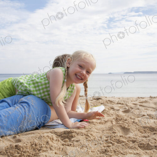A boy lying on his front on the sand looking out to sea. His sister lying on top of him. A boy lying on his front on the sand looking out to sea. His sister lying on top of him.