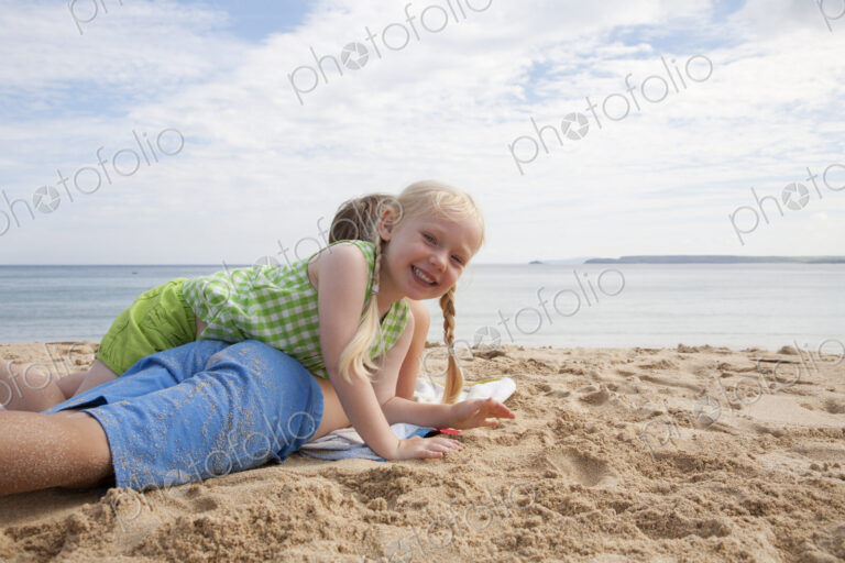 A boy lying on his front on the sand looking out to sea. His sister lying on top of him.