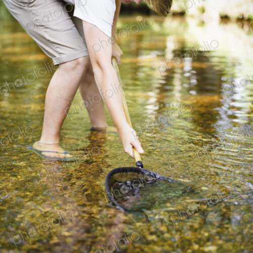 A boy paddling in shallow water, leaning over and using a small fishing net.
