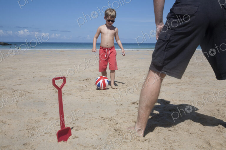 A boy playing football with a man on the sand. Father and son. A beach spade upright…