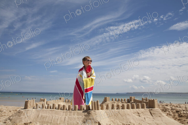 A boy standing beside a sandcastle, on top of a mound of sand. Beach.