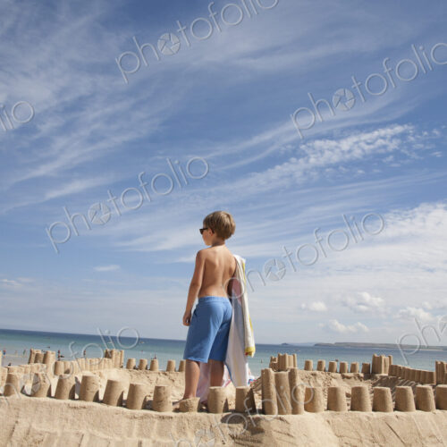 A boy standing beside a sandcastle, on top of a mound of sand. Beach. A boy standing beside a sandcastle, on top of a mound of sand. Beach.