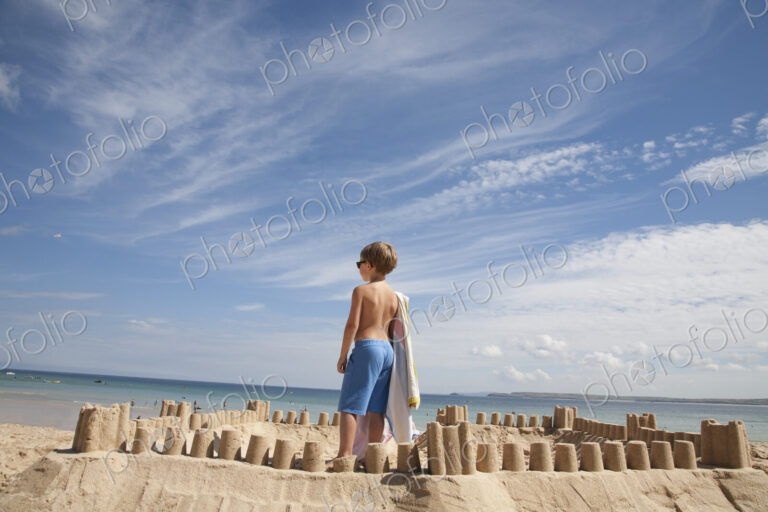 A boy standing beside a sandcastle, on top of a mound of sand. Beach.