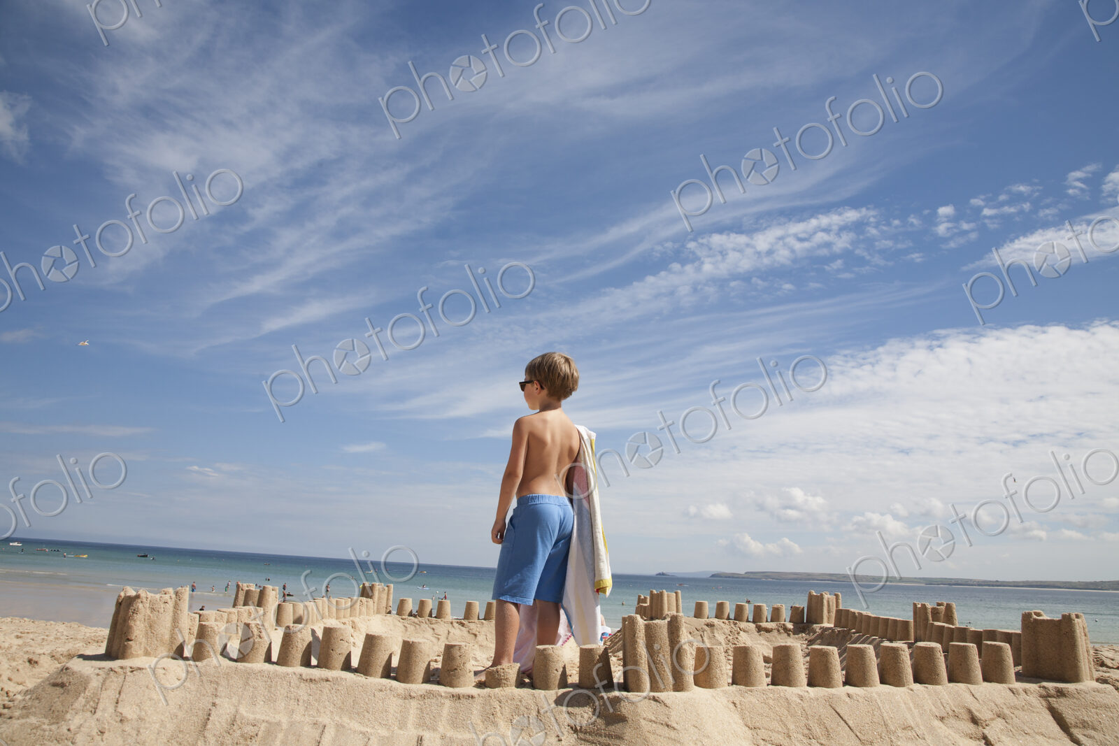 A boy standing beside a sandcastle, on top of a mound of sand. Beach.