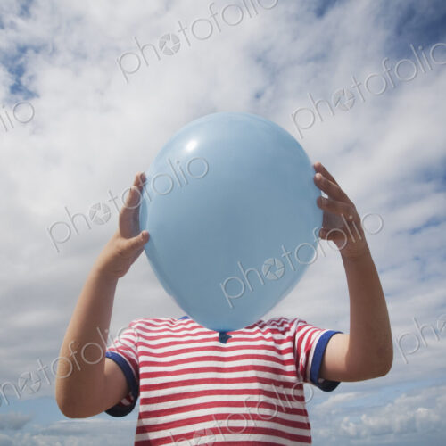 A boy standing holding a balloon in front of his face.