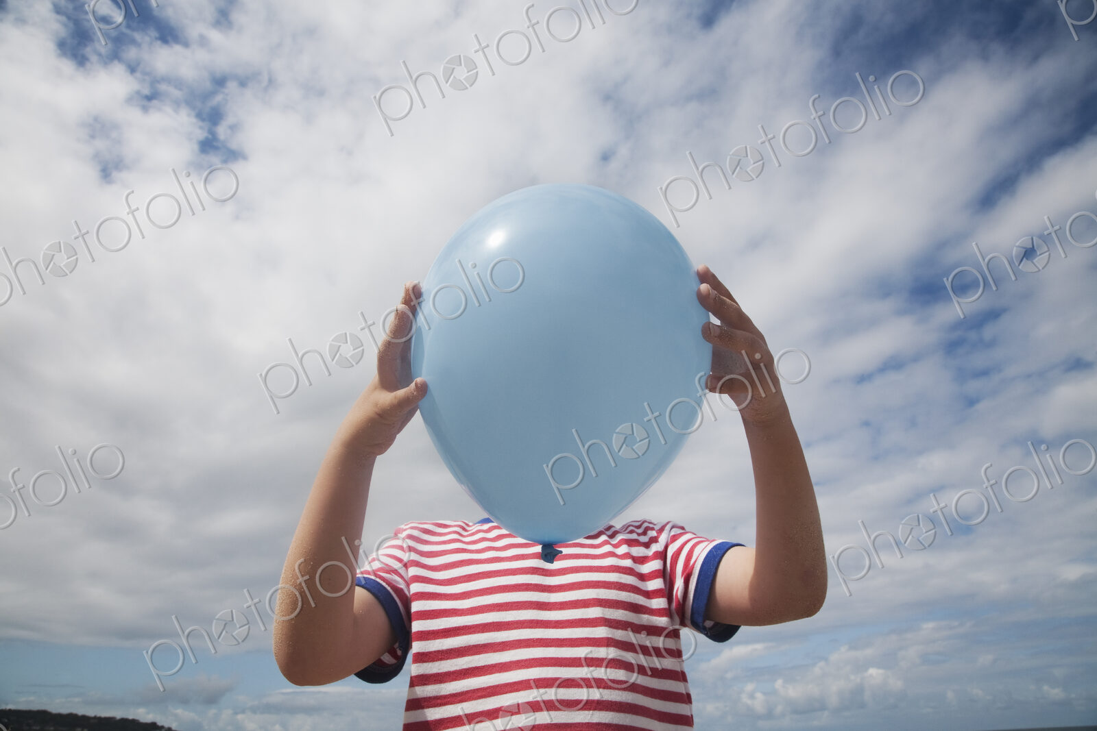 A boy standing holding a balloon in front of his face.