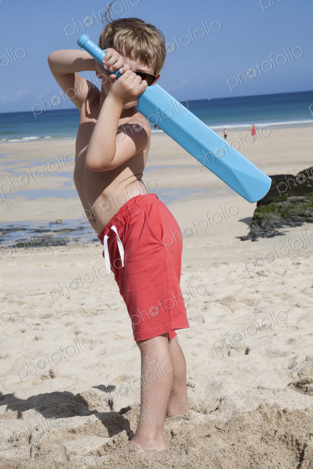 A boy standing on the sand with a small blue cricket bat in his hands.