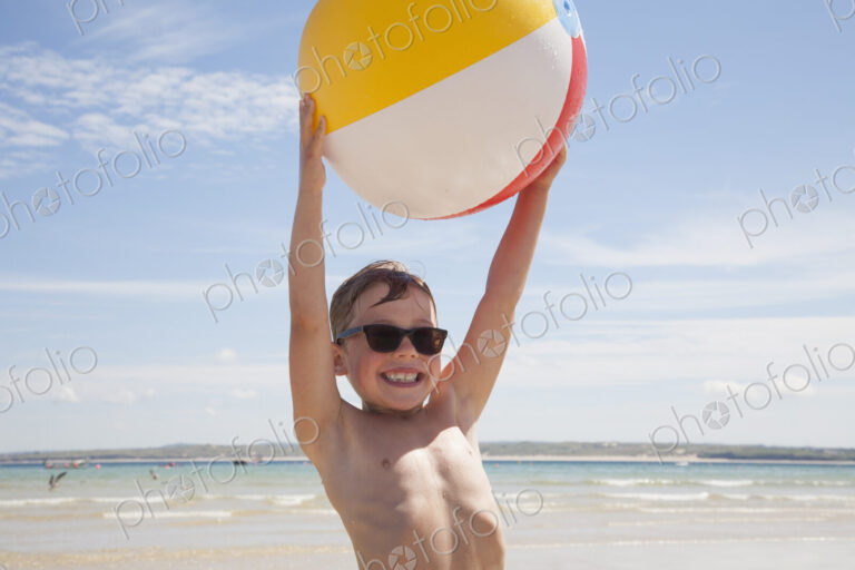 A boy wearing sunglasses on the beach. Holding an inflatable beach ball above his head.