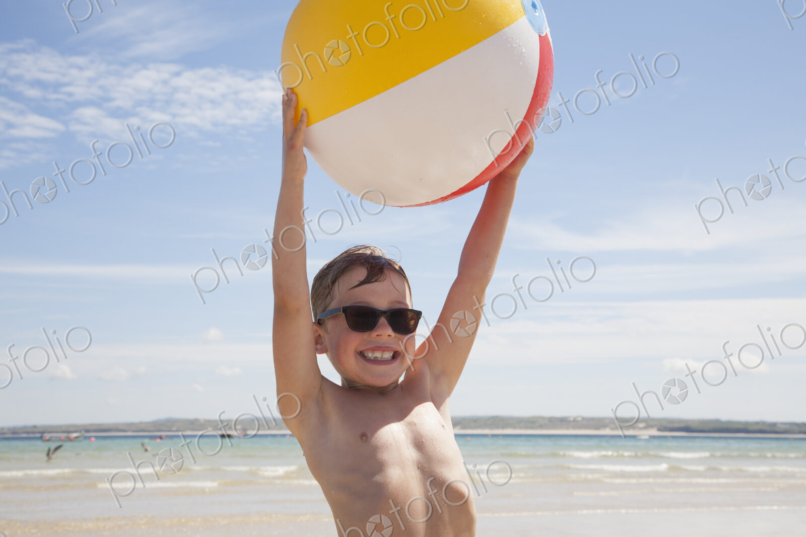 A boy wearing sunglasses on the beach. Holding an inflatable beach ball above his head.