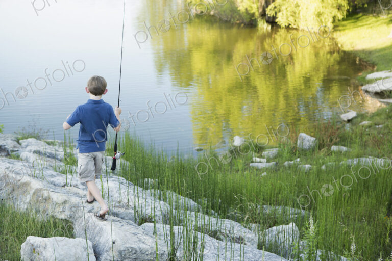 A boy with his fishing rod on the shore of a lake or river.