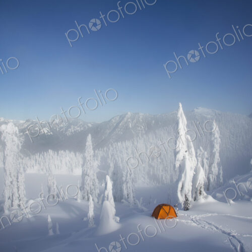 A bright orange tent among snow covered trees, on a snowy ridge overlooking a mountain… A bright orange tent among snow covered trees, on a snowy ridge overlooking a mountain…