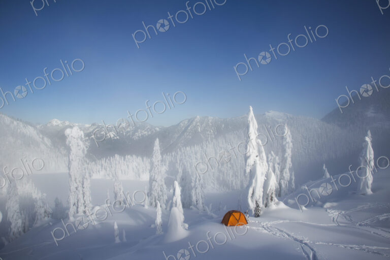 A bright orange tent among snow covered trees, on a snowy ridge overlooking a mountain…