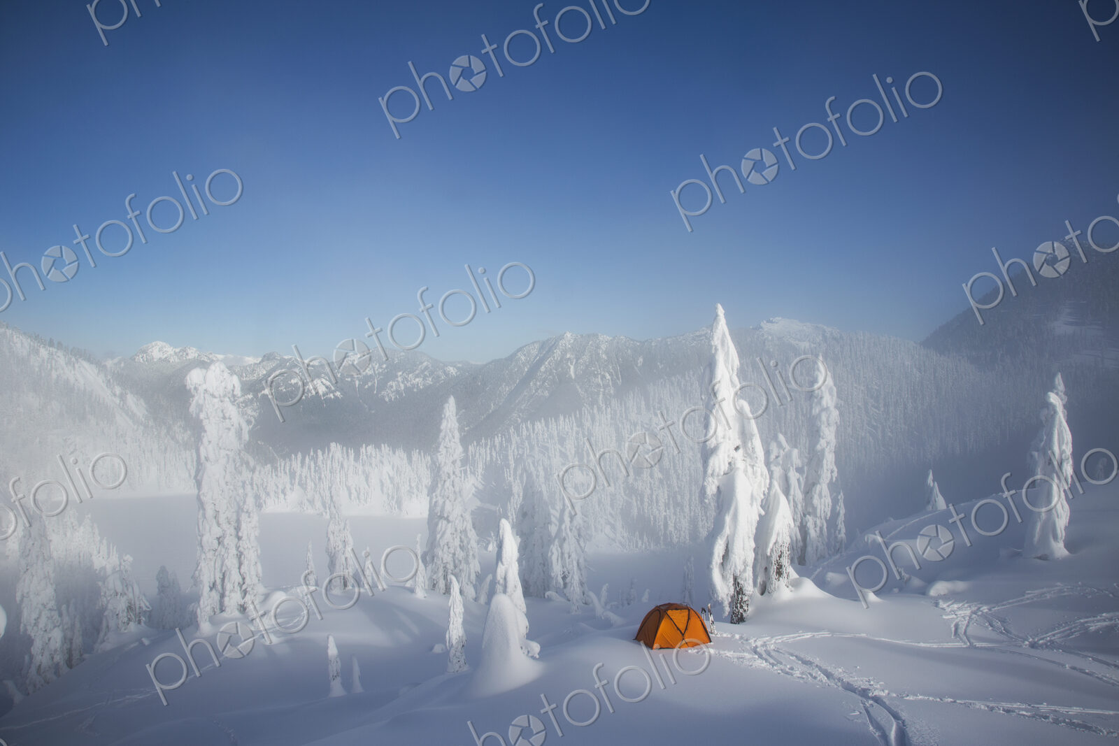 A bright orange tent among snow covered trees, on a snowy ridge overlooking a mountain…