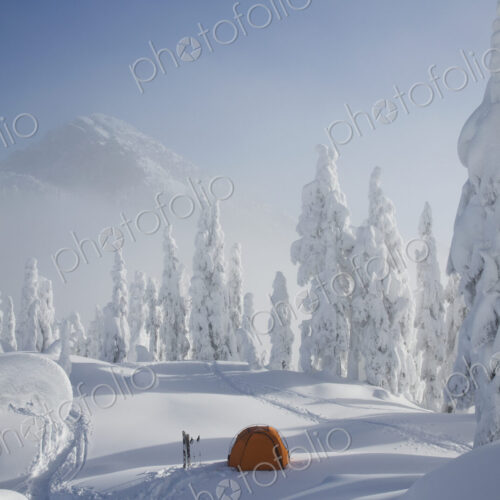 A bright orange tent sits on a snowy ridge overlooking a mountain in the distance. A bright orange tent sits on a snowy ridge overlooking a mountain in the distance.