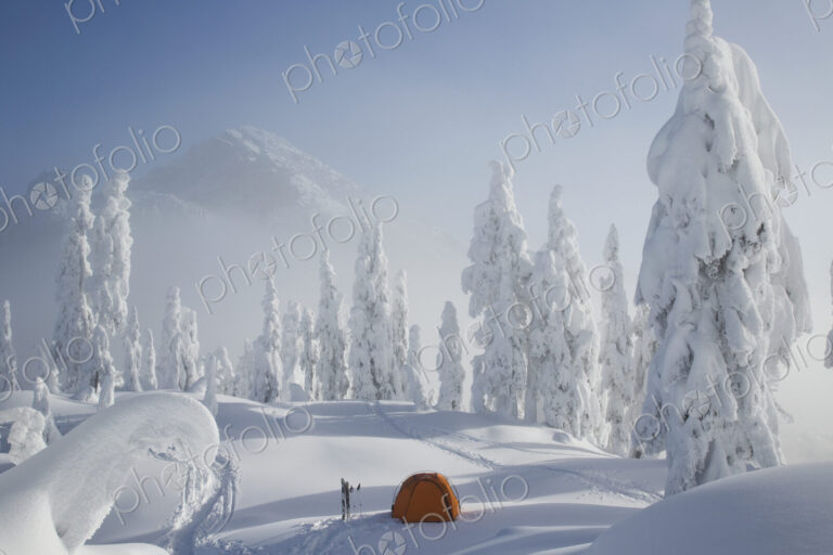 A bright orange tent sits on a snowy ridge overlooking a mountain in the distance.