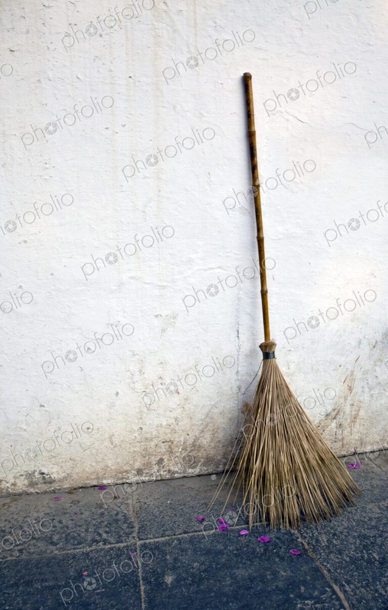 A broom made of twigs with a handle, for sweeping, leaning against a whitewashed wall.