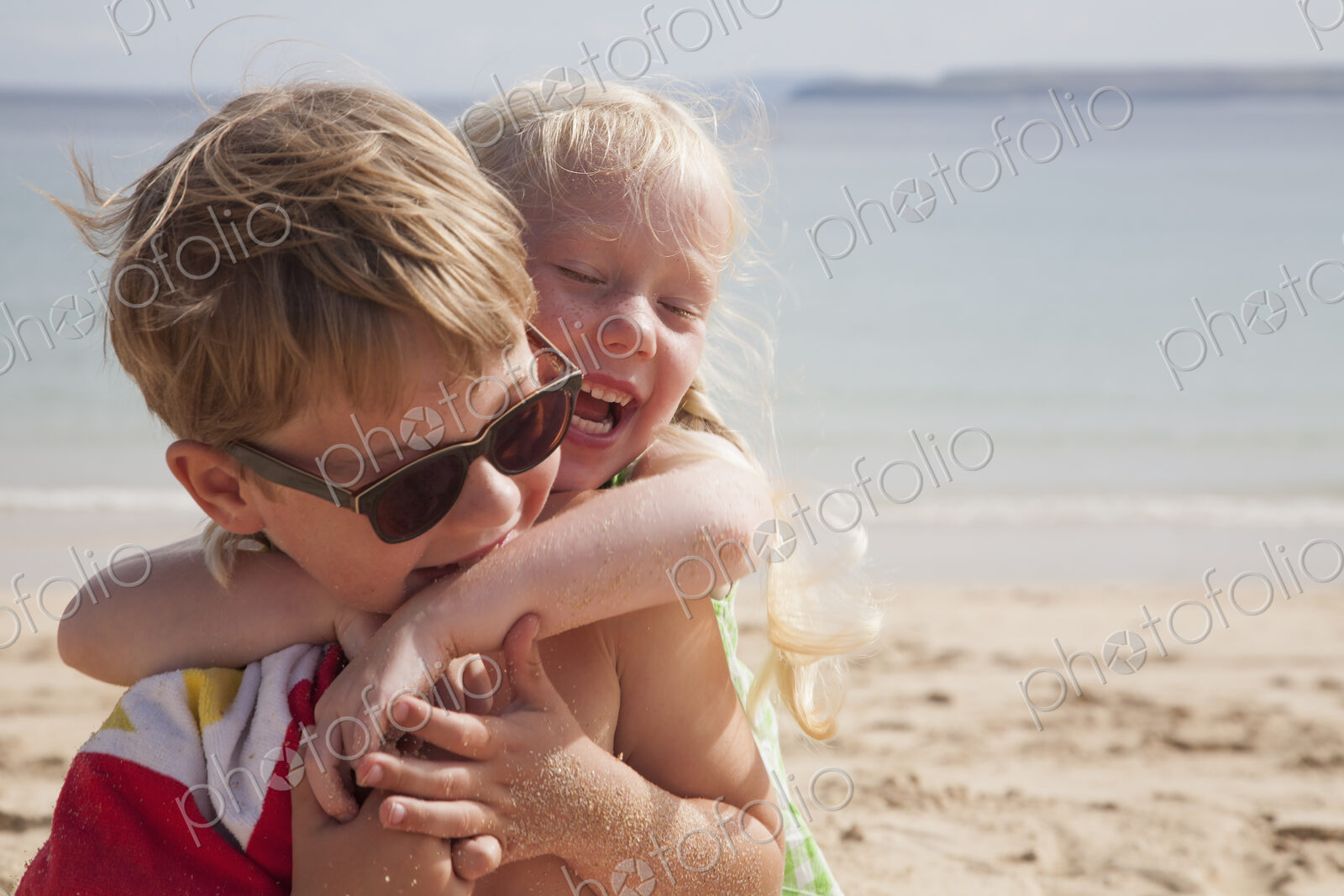 A brother and sister playfighting on the beach. A boy in sunglasses and a younger girl…