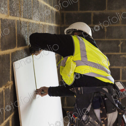 A builder in a hard hat wearing a tool belt, measuring a piece of wood.
