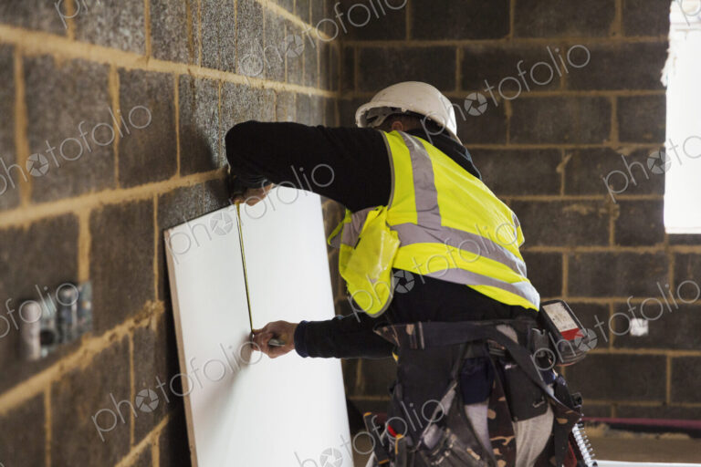 A builder in a hard hat wearing a tool belt, measuring a piece of wood.