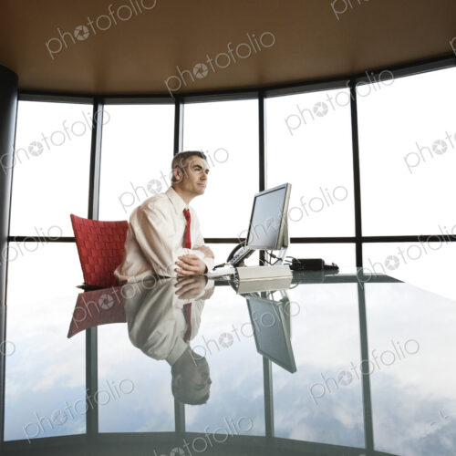 A businessman at his reflective office desk next to a large bank of windows. A businessman at his reflective office desk next to a large bank of windows.