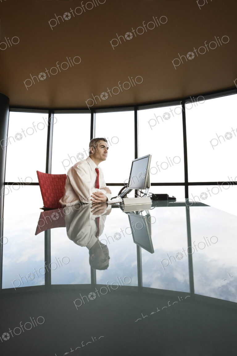 A businessman at his reflective office desk next to a large bank of windows.